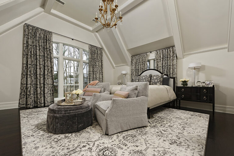 The high detailed ceilings adorned with a Visual Comfort chandelier in the master bedroom give you the feeling of being in a bed & breakfast. The large floral pattern on drapes grants balance to the dramatic ceilings. Cozy seating in front of the bed with custom chairs and ottoman for relaxing. INTERIOR DESIGN: MAX Interiors PHOTOGRAPHY: Bob Narod