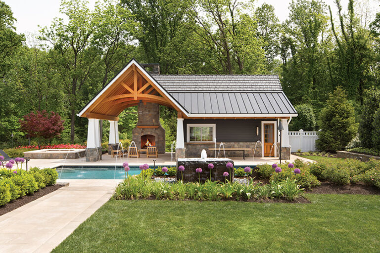 A Craftsman-style pool house features a stone fireplace and cedar beams adjacent to an expansive pool deck and a custom granite water feature. LANDSCAPE ARCHITECTURE: Surrounds Landscape Architecture + Construction PHOTOGRAPHY: Morgan Howarth