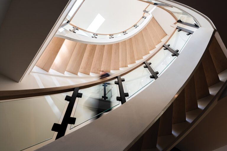 A sculptural, open-riser stair became the centerpiece of a Potomac abode, with wood treads and a glass railing accented in dark metal; a skylight fills the stairwell with natural light.