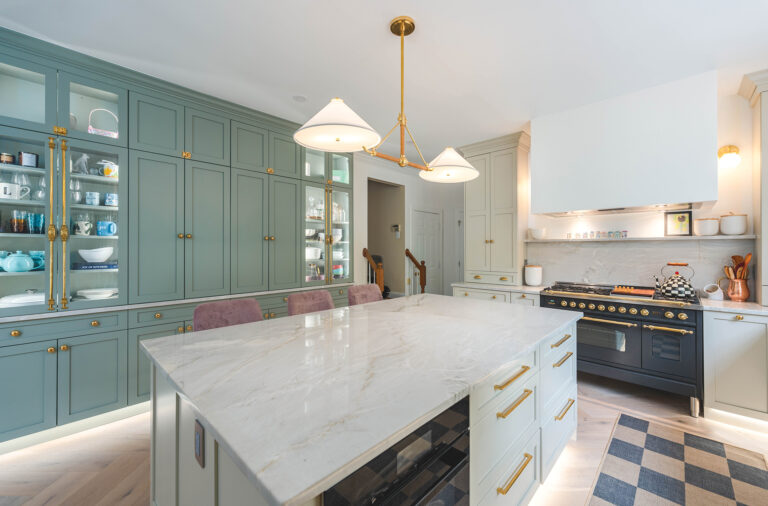 In a Great Falls kitchen, custom, sage-green cabinets and brass accents surround a quartzite-topped island. Wood floors in a herringbone pattern ground the space.